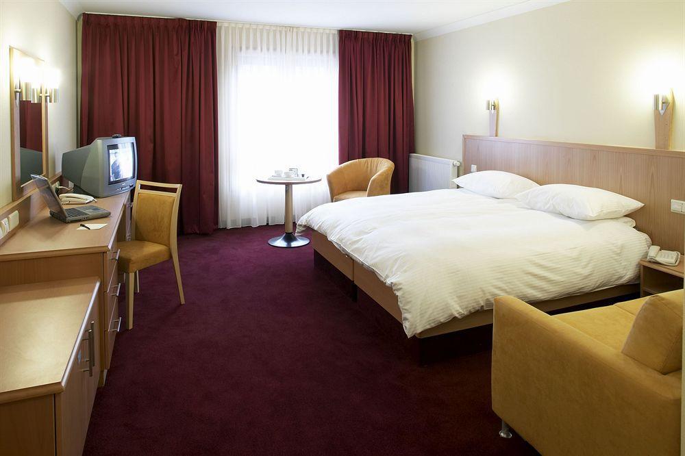 Clayton Hotel Dublin Airport Coolock Ruang foto The photo shows a hotel room featuring a double bed with white linens and pillows. There are two bedside lamps mounted on the wall above the bed. To one side, there's a small round table with two chairs. Opposite the bed, a wooden desk with a chair i